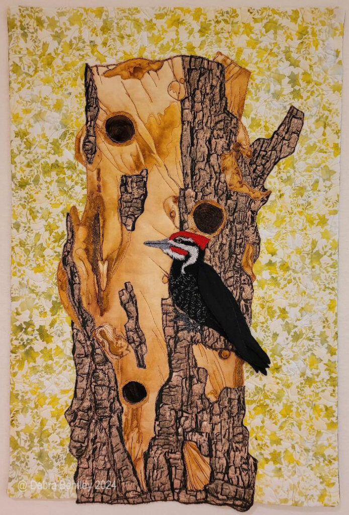 Woodpecker Snag My art quilts express my wonder at the beauty and tenacity of nature. Fabric and thread capture colors and textures, lights and shadows, emotions of joy and sorrow. The common thread through all my pieces is the delicate balance between humankind and nature, and the sense of spiritual renewal that oneness brings. This quilt is about how even decaying and dying trees and plants are essential parts of the life cycle of animals and birds. Woodpeckers and other birds and animals, often rely on dead and dying trees as a source of insects for food and cavities for shelter. I used rust dyed fabric and bark printed fabric for the tree, adding free motion stitching. The woodpecker is based on photos I’ve taken around our feeders and trees. He is appliqued on the tree. The bird is free-motion embroidered, with some of his wing feathers stitched onto him.